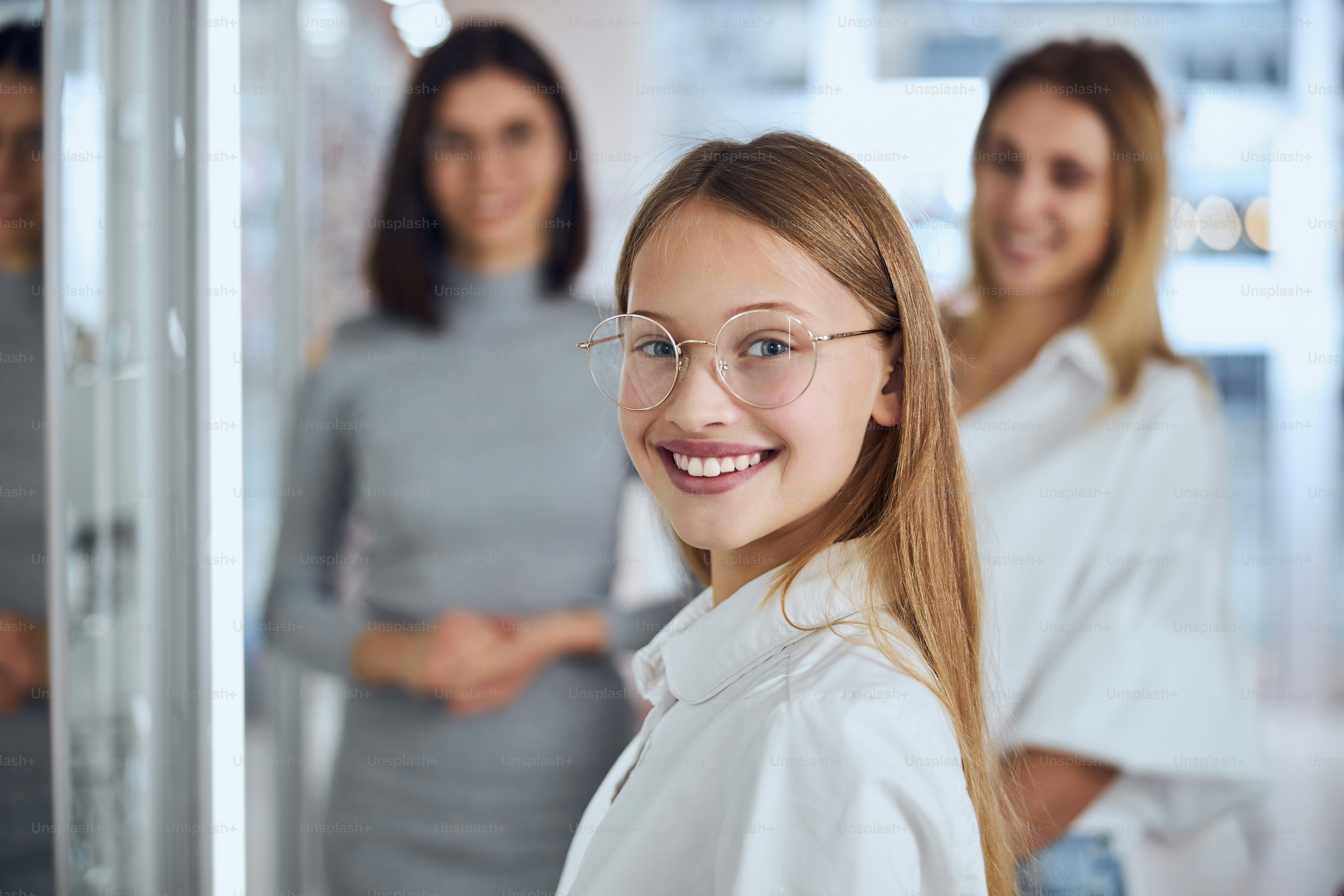 Close up side view portrait of happy cheerful teenager girl in white shirt and eyeglasses looking and posing at the photo camera in ophthalmology clinic
