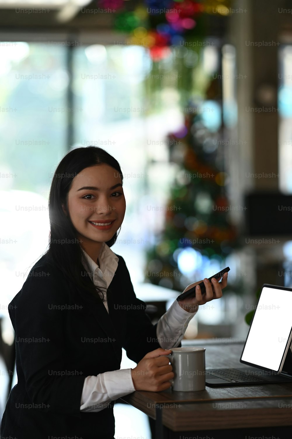 Portrait d’une femme d’affaires souriante assise sur son lieu de travail tenant un téléphone intelligent et souriant à la caméra.