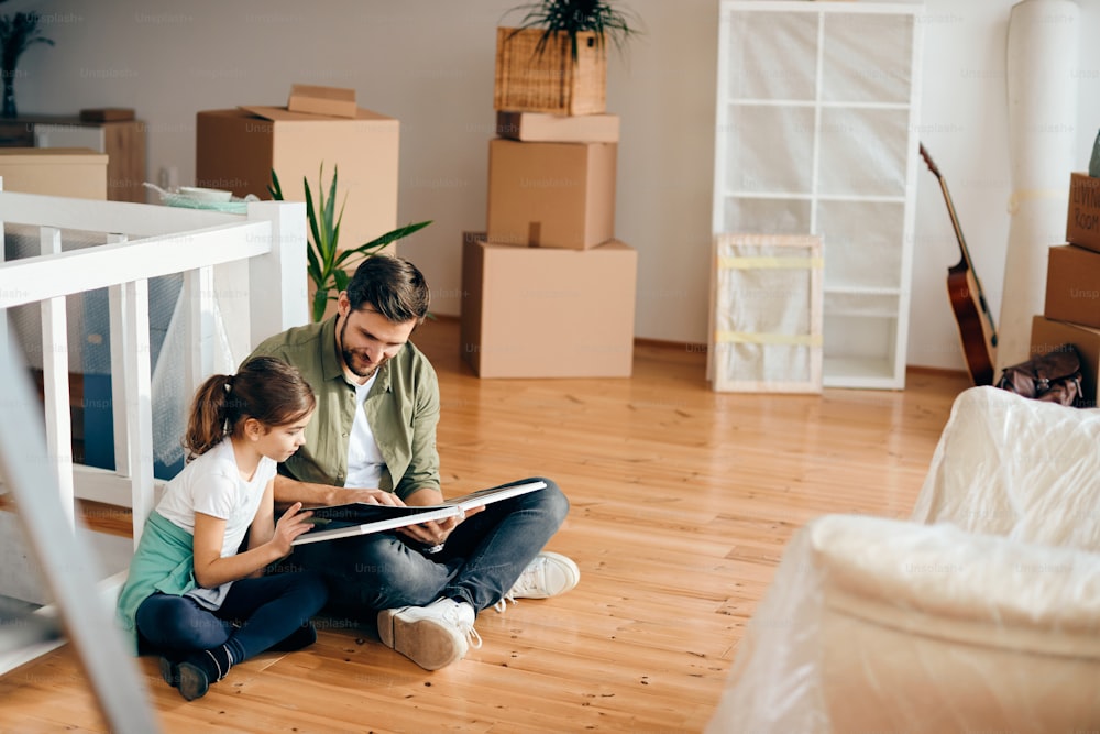 Smiling father and daughter reading book while sitting on the floor at their new house.