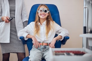 Bella ragazza adolescente in posa e guardando la fotocamera mentre è seduta sul sedile con cornici di prova sul viso nel moderno centro oftalmologico