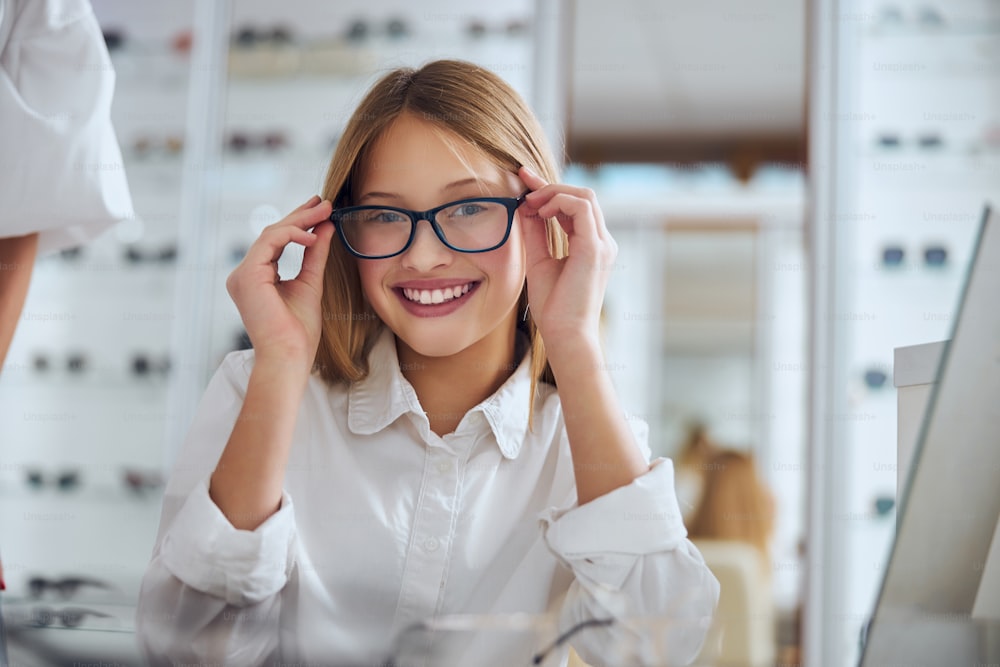 Close up portrait of happy cheerful female child wearing eyeglasses while sitting in optician center