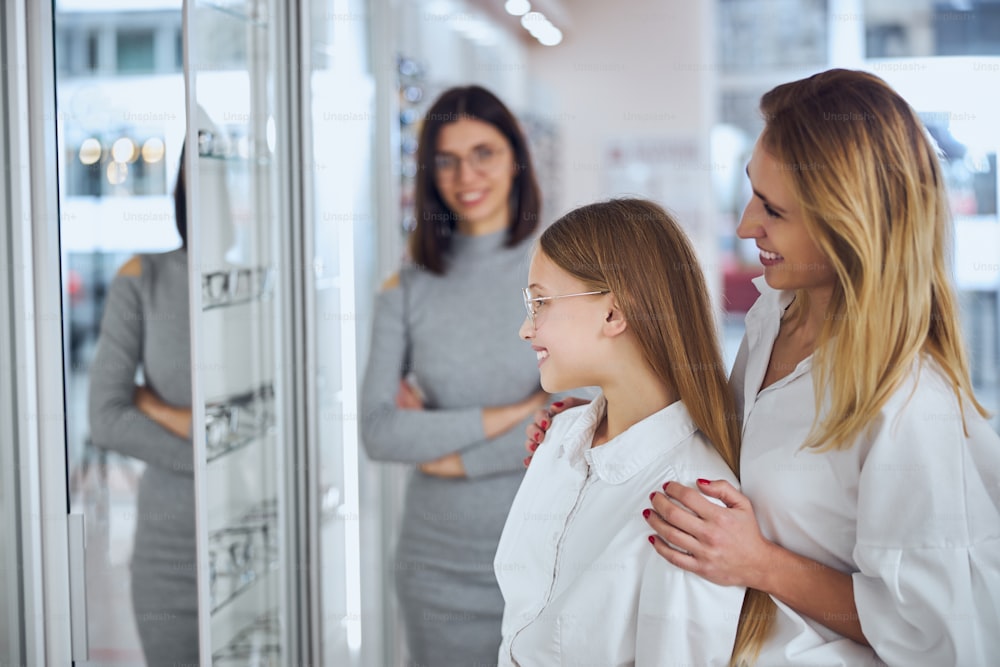 Portrait en gros plan de vue latérale d’une mère et d’une fille heureuses et souriantes, choisissant et essayant des lunettes dans un magasin d’optique