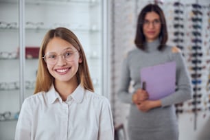 Waist up portrait of pretty elegant teenager girl in white shirt standing in front of the woman in grey dress in room indoors