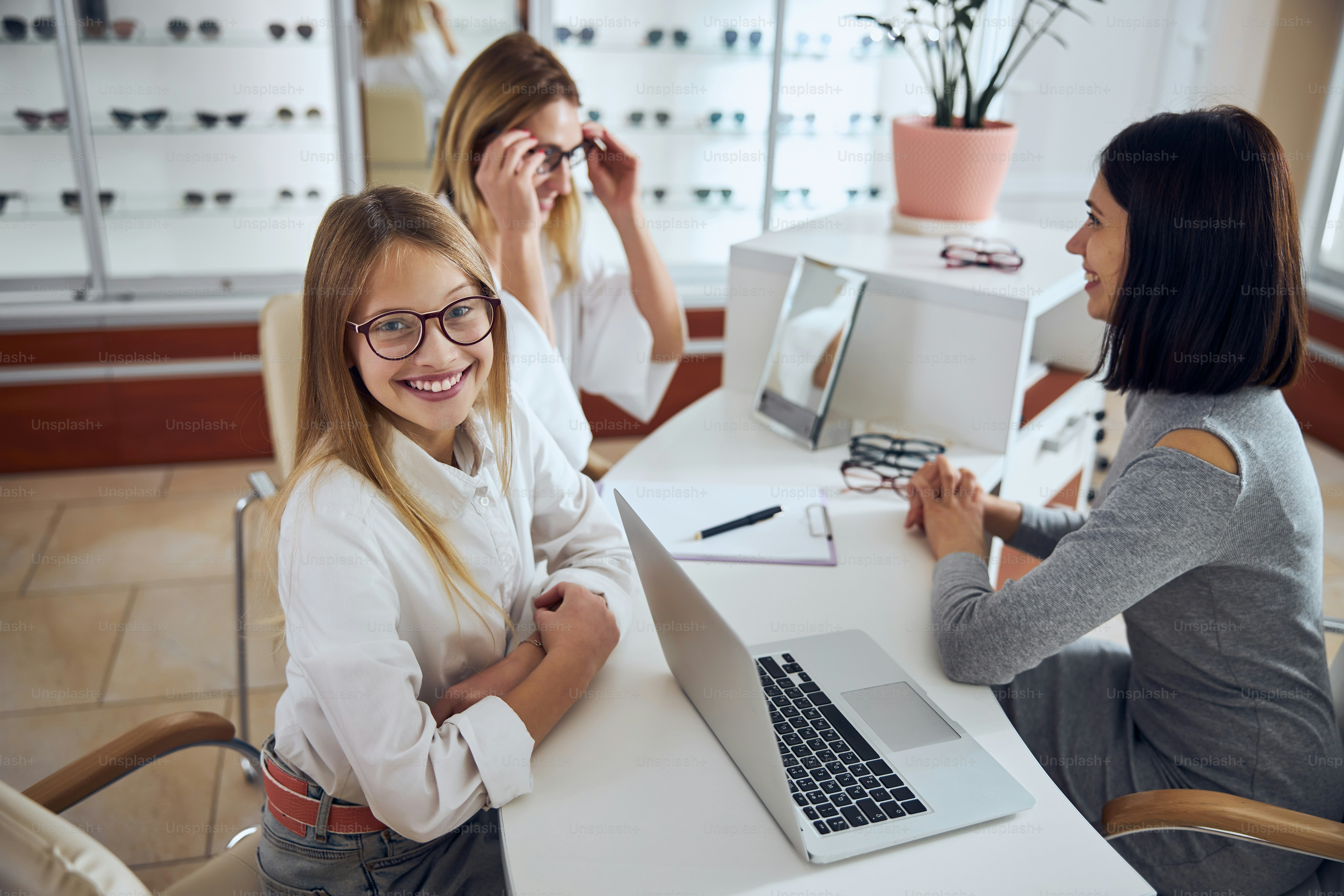 Fmale child sitting looking at the photo camera in new glass while her mother choosing glass for herself in office optician