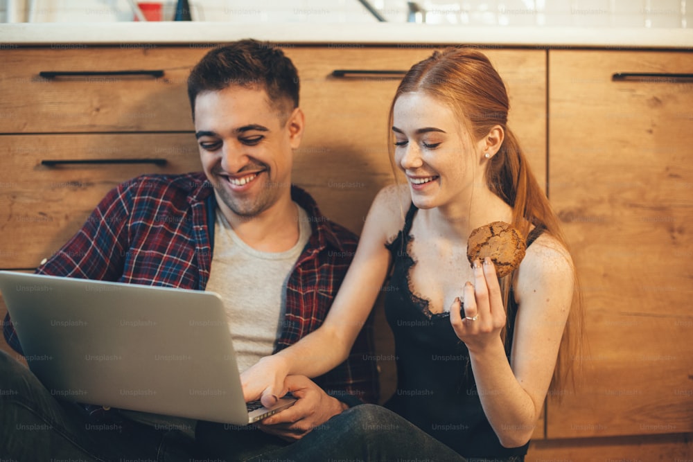 Caucasian man with his wife laughing in the kitchen on the floor using a laptop and eating cookies