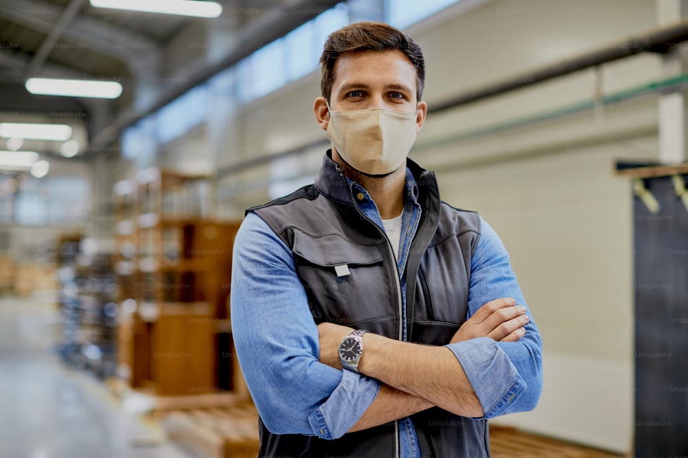 Ingeniero carpintero con mascarilla mientras está de pie con los brazos cruzados en una fábrica y mira a la cámara.