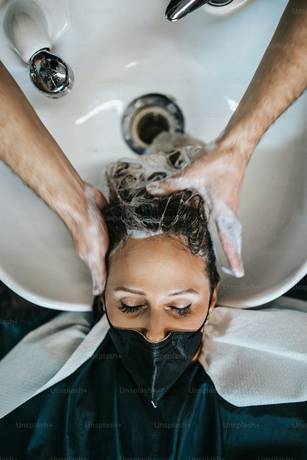 Peluquero lavando el cabello de una hermosa mujer adulta joven en la peluquería. Lleva una mascarilla protectora como protección contra la pandemia del virus.
