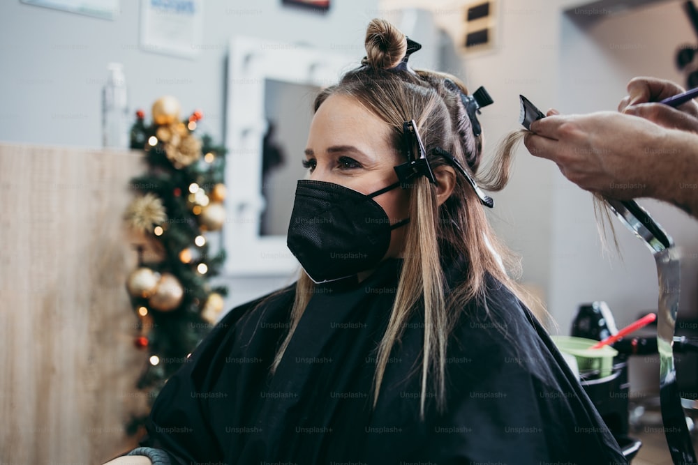 Il parrucchiere tinge i capelli femminili, mettendo in evidenza i capelli al suo cliente con una pellicola. Indossano una maschera protettiva per il viso come protezione contro la pandemia di virus.