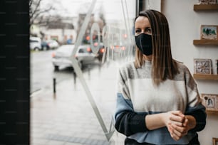 Junge Künstlerin steht vor Werkstattfenstern oder Türen und schaut nach draußen. Sie trägt eine schützende Gesichtsmaske. Kunst im Coronavirus Lockdown-Zeitkonzept.