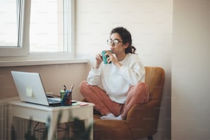Photo of a concentrated woman with glasses drinking tea at home sitting in an armchair and watching something at the laptop