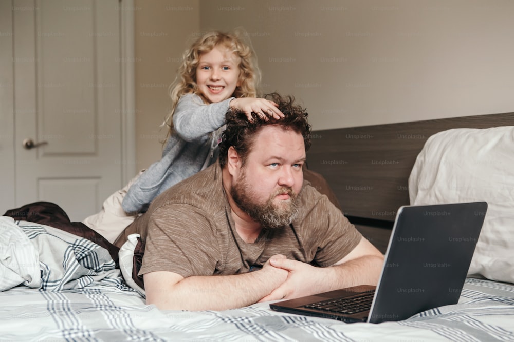 Trabaja desde casa con niños. Padre trabajando en una computadora portátil en el dormitorio con una hija pequeña en su espalda. Momento familiar divertido y sincero. Nueva normalidad durante el confinamiento por la cuarentena por el coronavirus.