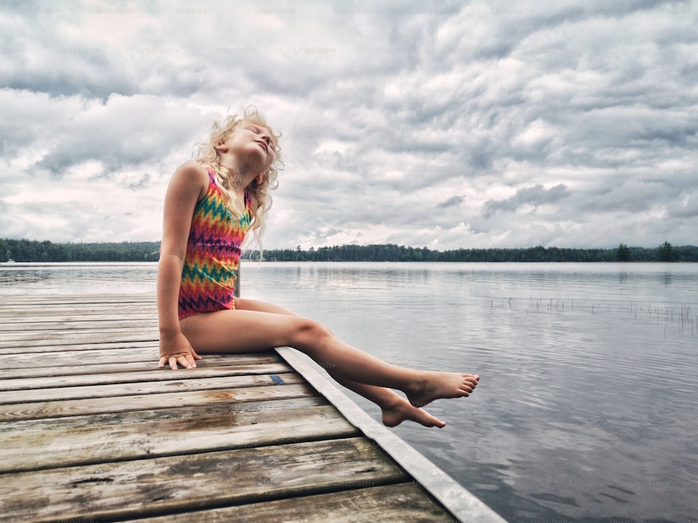 Cute adorable blonde Caucasian girl sitting on wooden dock by lake and looking up in the sky. Dreaming relaxing cute child kid by water outdoor on summer day. Countryside slow life living.