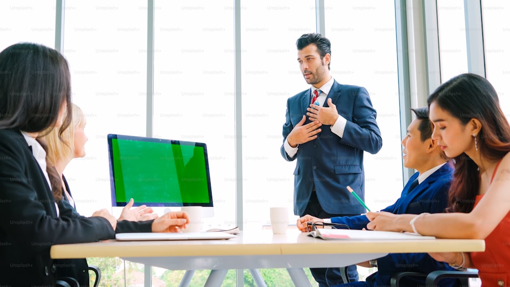 オフィスのテーブルの上にグリーンスクリーンのクロマキーテレビやコンピューターがある会議室のビジネスマン。ビデオ会議通話で会議中のビジネスマンとビジネスウーマンの多様なグループ。