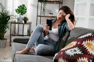 Sad woman sitting on the couch, using the phone. Upset woman waiting for a phone call.