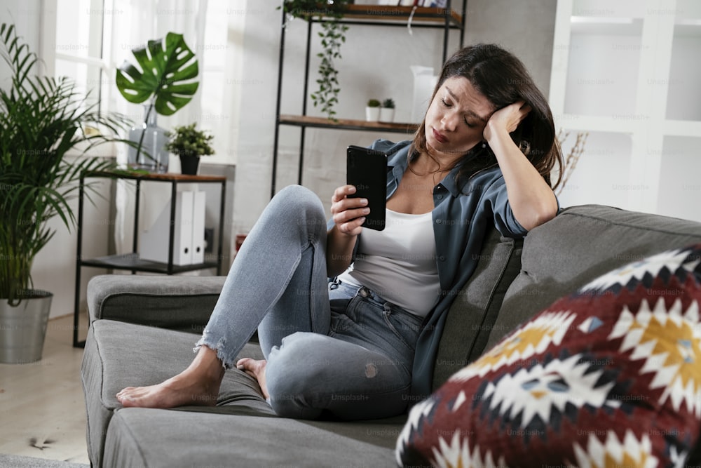 Donna triste seduta sul divano, usando il telefono. Donna sconvolta in attesa di una telefonata.