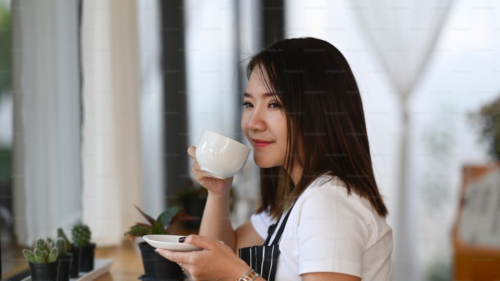 Vista laterale della donna barista asiatica che tiene una tazza di caffè e annusa nella caffetteria.