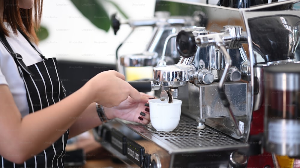 Vista de cerca de una joven barista con delantal haciendo café usando una cafetera en la cafetería.