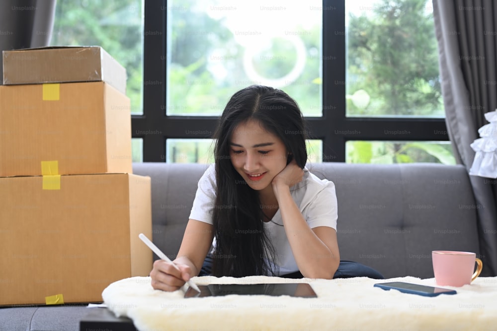 디지털 태블릿으로 작업하고 집에서 상자에 둘러싸여 앉아 있는 중소기업 기업가 여성을 시작합니다.