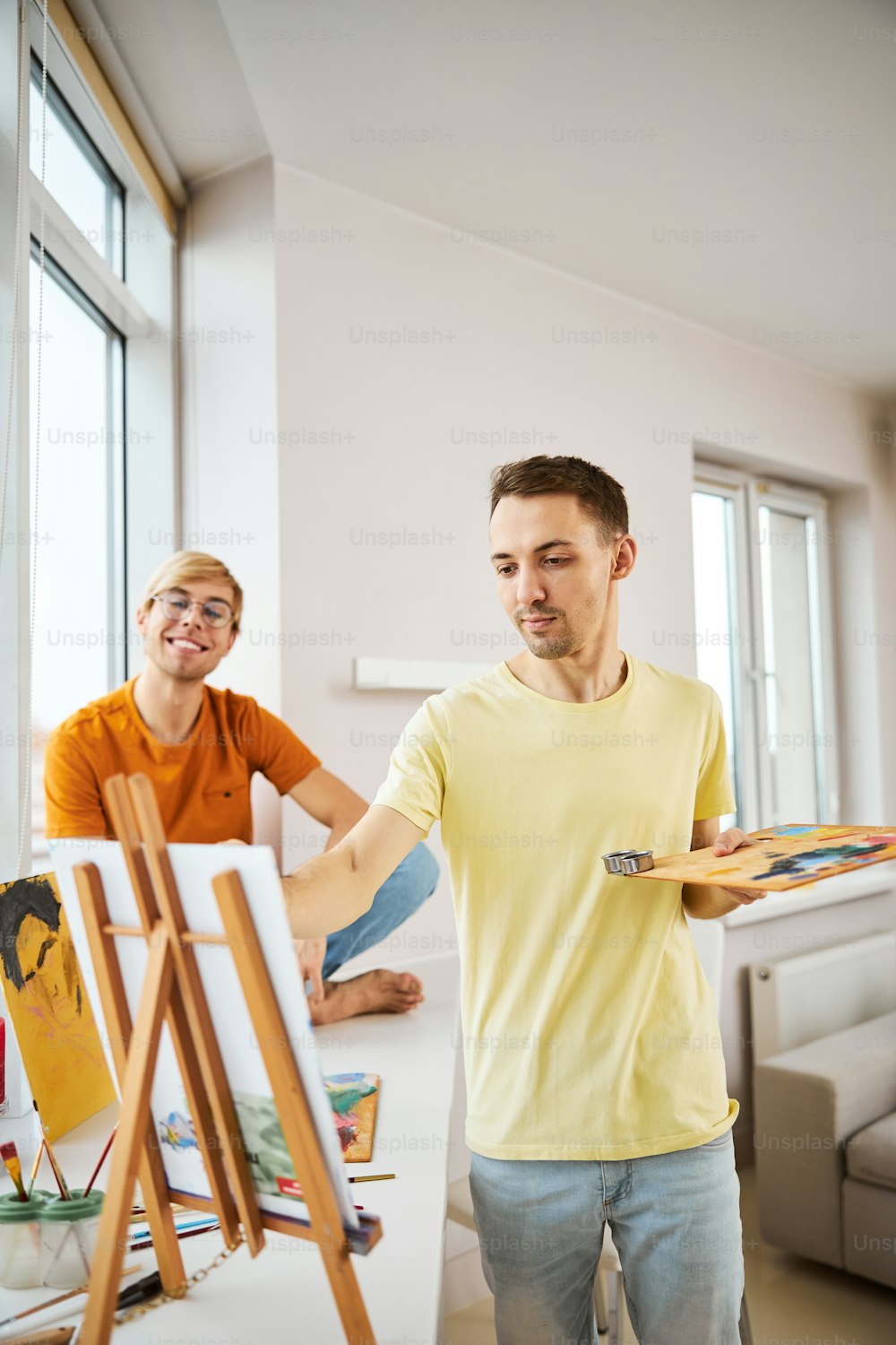 Handsome male artist painting on canvas with acrylic paints while his boyfriend sitting on windowsill and smiling