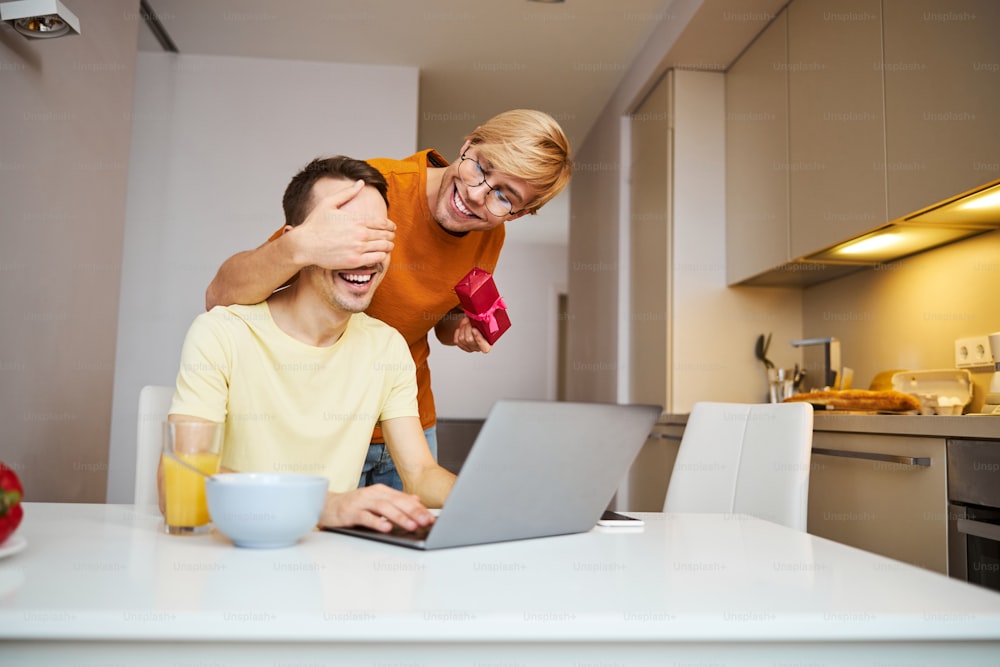 Handsome young man using laptop and smiling while joyful boyfriend holding gift box