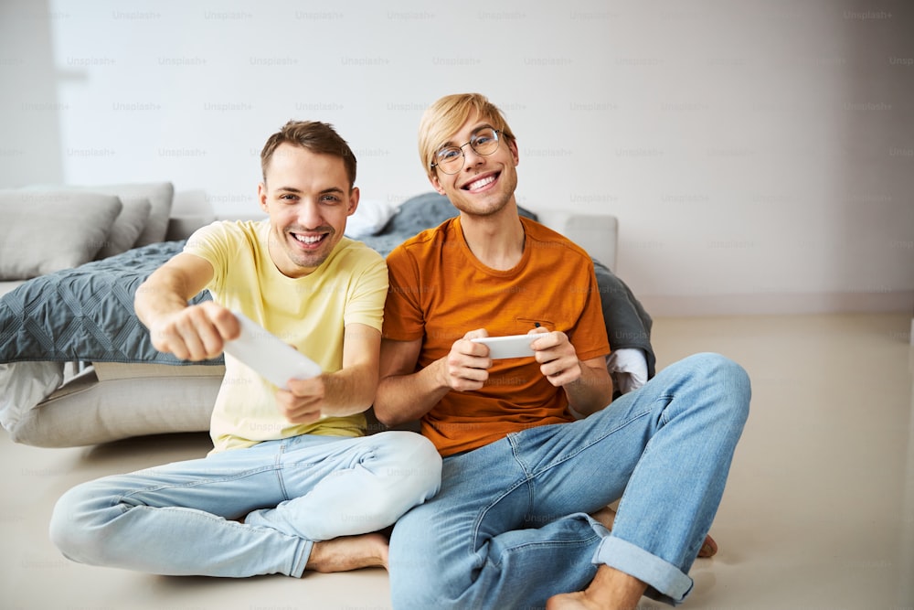 Handsome cheerful guys looking at camera and smiling while using smartphones as joystick controllers
