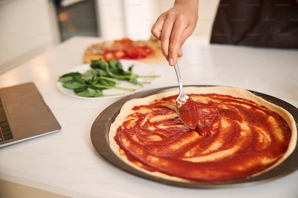 Hand with tea spoon spreading tomato sauce on the surface of a pizza base on a baking pan