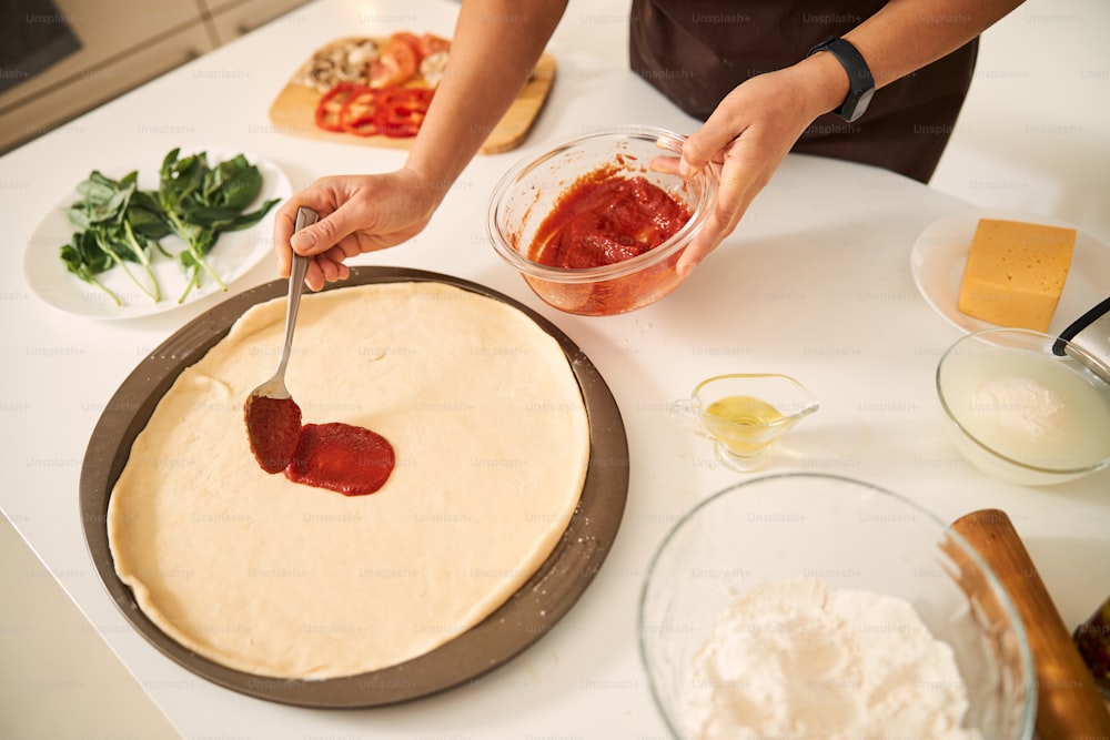 Close up of a woman holding a bowl of tomato sauce and spreading it on the pizza base