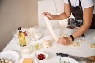 Woman holding a smooth piece of dough above the table while standing near the pasta machine in the kitchen