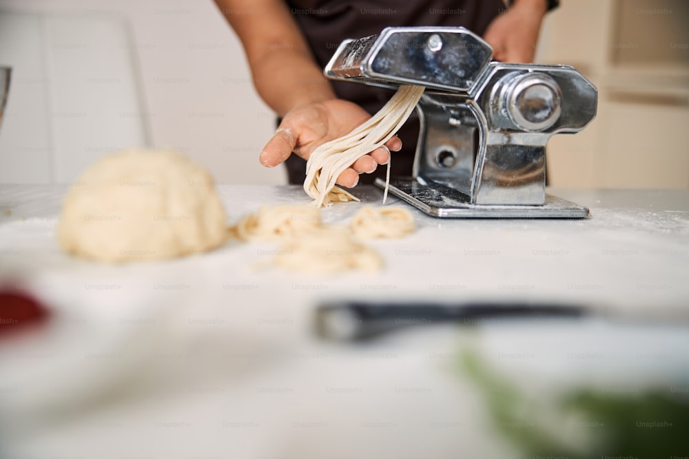 Close up photo of hands of unrecognized person taking stripes of dough from the pasta machine in the kitchen