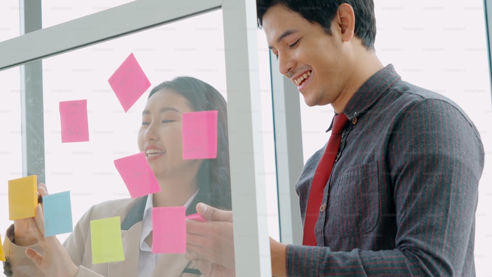 Business people work on project planning board in office and having conversation with coworker friend to analyze project development . They use sticky notes posted on glass wall to make it organized .