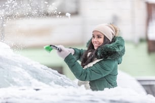 Fêmea jovem em roupas de inverno limpa o carro da neve fora do bloco de apartamentos no inverno.