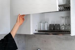 Vista recortada de la mujer abierta a mano armarios blancos con tazas, tazas y cristalería, de pie en la cocina moderna. Concepto de costoso mecanismo de elevación en muebles
