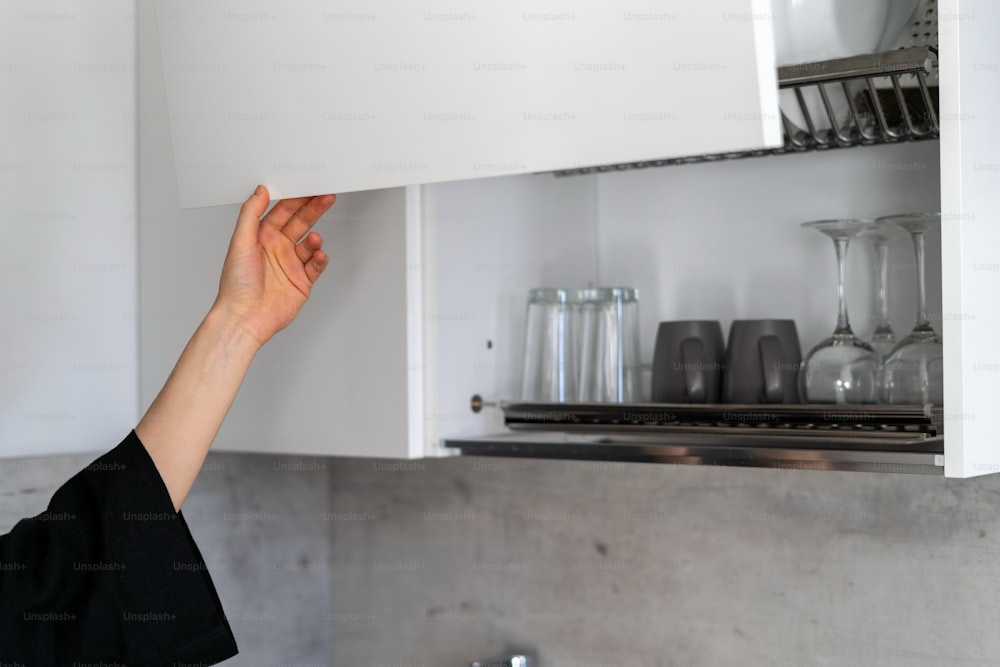 Vista ritagliata di armadi bianchi aperti a mano della donna con tazze, tazze e bicchieri, in piedi sulla cucina moderna. Concetto di costoso meccanismo di sollevamento nei mobili