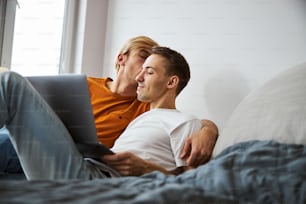 Handsome young man watching movie on notebook and smiling while loving boyfriend kissing his cheek