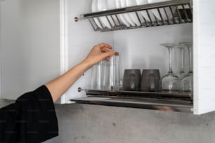 Cropped view of woman hand taking clean glassware from shelves. Housewife standing on modern kitchen near open white cupboards full of kitchenware
