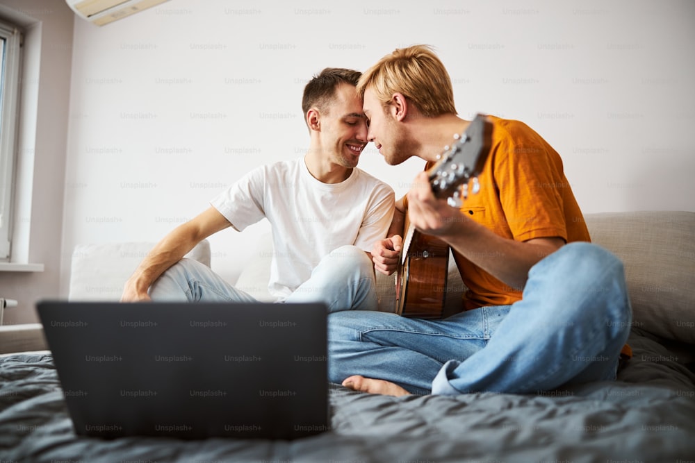 Handsome young man holding guitar and sharing tender moment with boyfriend while spending time at home