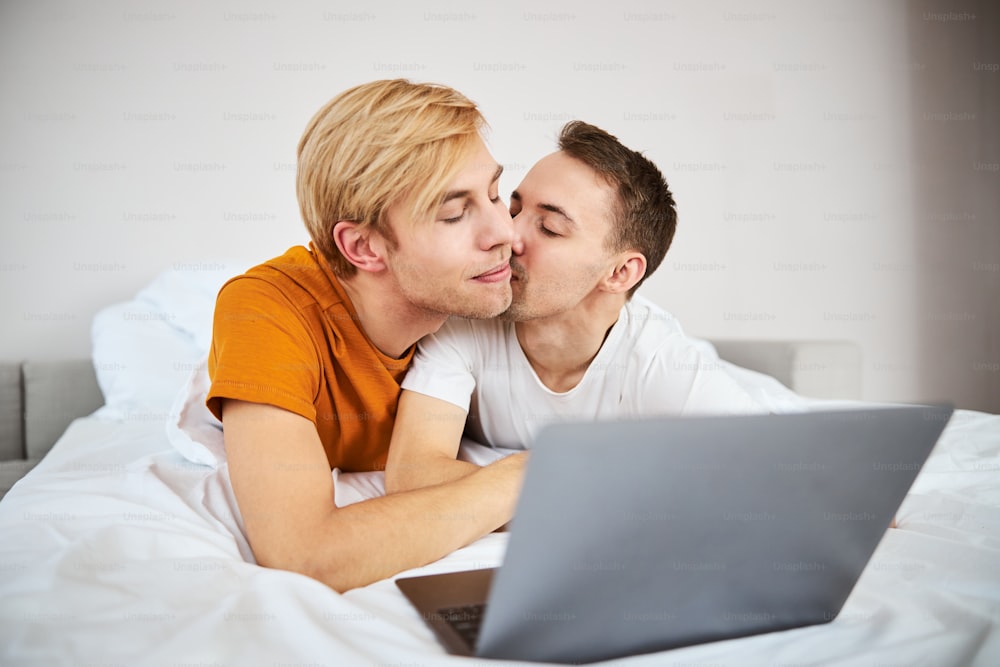 잘생긴 청년이 침대에 누워 노트북에 영화를 보고 있는 동안 남자친구가 그의 뺨에 키스