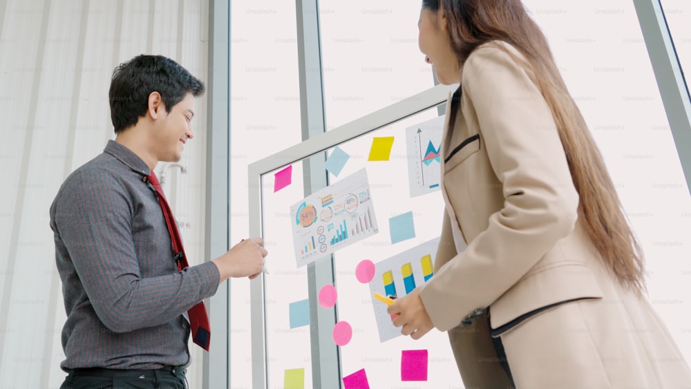 Business people work on project planning board in office and having conversation with coworker friend to analyze project development . They use sticky notes posted on glass wall to make it organized .