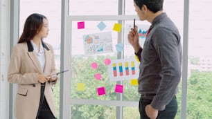 Business people work on project planning board in office and having conversation with coworker friend to analyze project development . They use sticky notes posted on glass wall to make it organized .