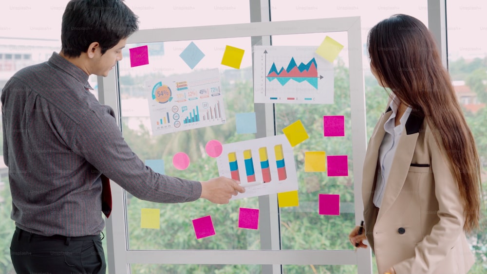 Business people work on project planning board in office and having conversation with coworker friend to analyze project development . They use sticky notes posted on glass wall to make it organized .