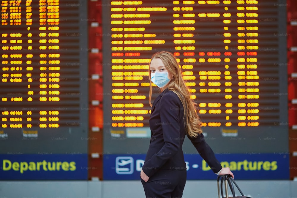 Woman wearing protective face mask with hand luggage in international airport terminal, looking at information board, checking her flight. Cabin crew member with suitcase. Traveling during pandemic