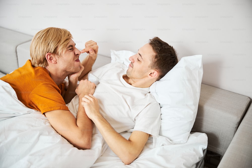 Joven guapo tocando la nariz de su novio y sonriendo mientras descansa sobre sábanas blancas en casa