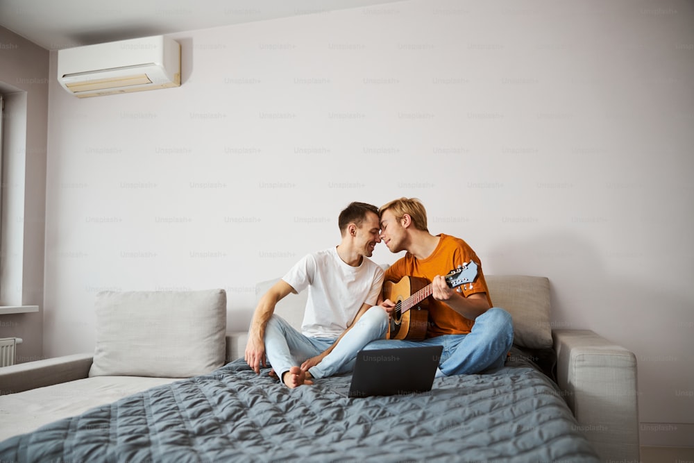 Handsome young man playing guitar and sharing tender moment with boyfriend while spending time at home