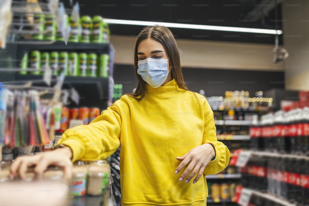 Donna che indossa una maschera protettiva e compra cibo in un negozio di alimentari durante l'epidemia di virus. giovane donna che indossa una maschera protettiva e guanti che fa la spesa in un periodo di pandemia di virus, acquistando forniture alimentari