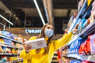 Distanciation sociale dans un supermarché. Une jeune femme portant un masque jetable achète de la nourriture et la met dans un panier d’épicerie. Shopping pendant l’épidémie de Coronavirus Covid-19 2020
