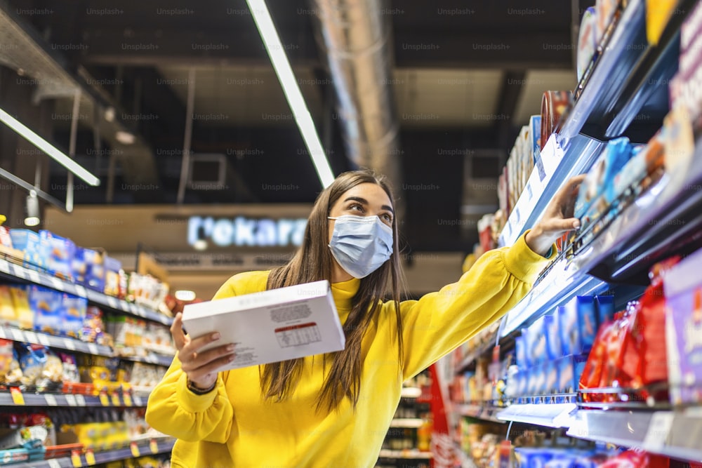 スーパーマーケットでのソーシャルディスタンス。使い捨てのマスクを着けた若い女性が食べ物を買ってきて、買い物かごに入れている。コロナウイルスCovid-19流行時のショッピング2020