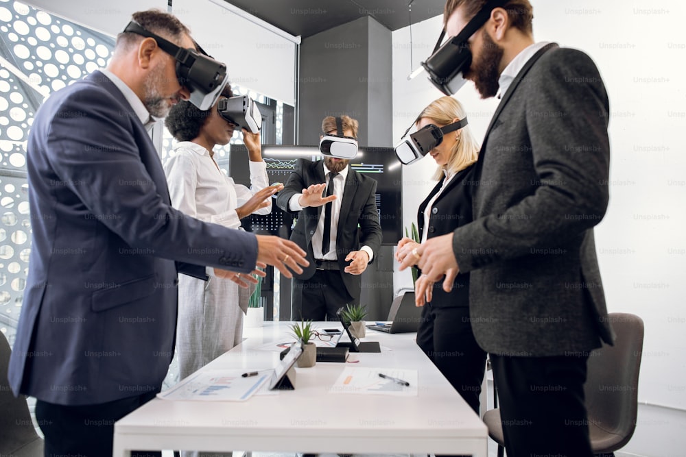 Team von kreativen multirassischen Kollegen, Designern, Architekten in formeller Kleidung, die um den Tisch stehen und eine VR-Brille zur Visualisierung des Projekts verwenden. Konzept der virtuellen 3D-Technologien.