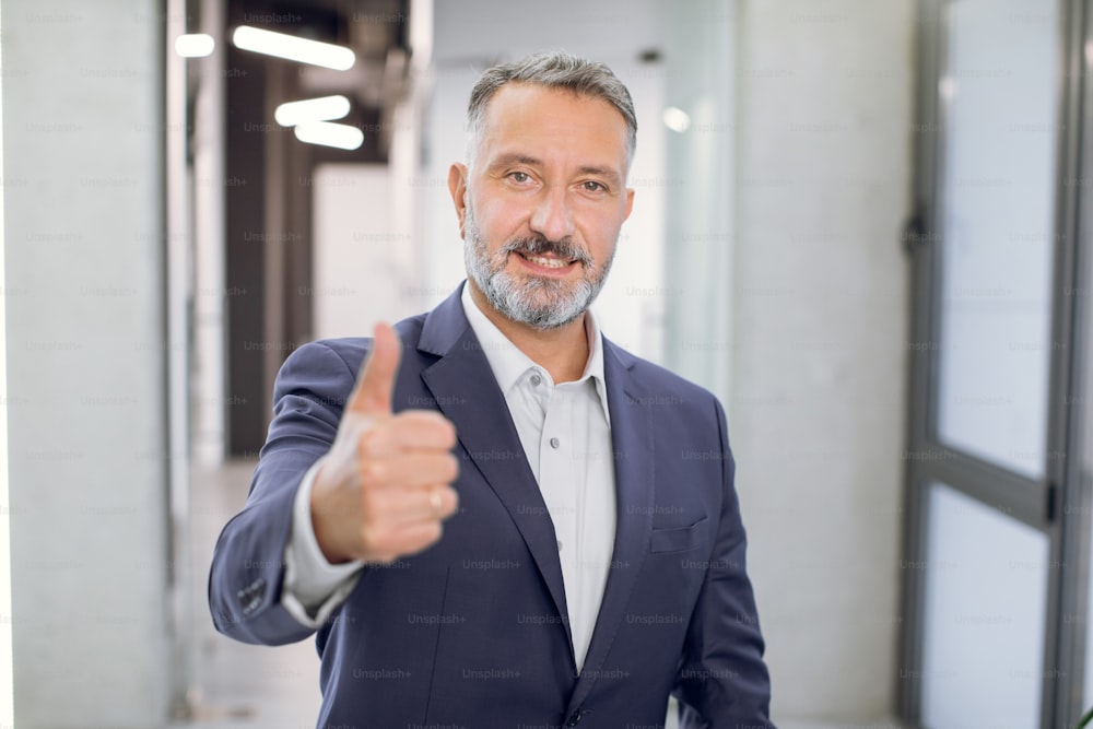 Cheerful handsome middle aged bearded Caucasian businessman, wearing shirt and jacket, showing thumbs up, posing and smiling at camera, in front of modern light loft office corridor.