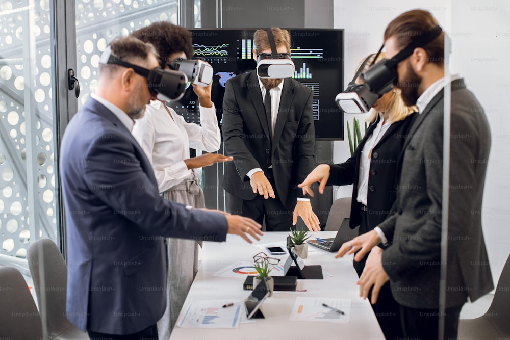 Équipe de collègues multiraciaux créatifs, designers, architecte en tenue de soirée, debout autour de la table et utilisant des lunettes VR pour la visualisation du projet. Concept de technologies 3D virtuelles.