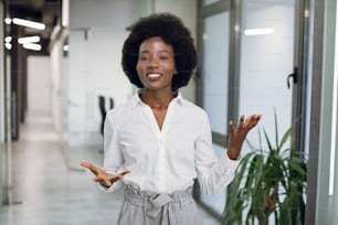 Young attractive and successful black afro american business woman in formal wear, giving an interview, speaking and gesturing, while standing indoors in modern office building.